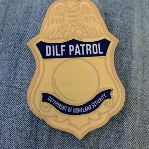 May include: A gold-colored badge with a blue ribbon that says "DILF PATROL" and "DEPARTMENT OF HOMELAND SECURITY".