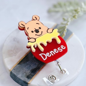 May include: A red heart-shaped Winnie the Pooh themed retractable badge holder with a yellow honey pot and the name "Denese" written on it.
