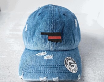 May include: Distressed denim baseball cap with a patch featuring the Oklahoma state outline in black with a red stripe. The cap has a curved brim and a circular logo on the side.