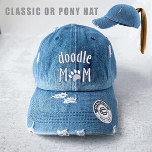 Puede incluir: Gorra de béisbol de mezclilla desgastada con el texto "doodle M M" bordado en blanco. La gorra tiene una visera curva y una abertura para cola de caballo. El diseño incluye una huella de pata y un logotipo en el lateral.