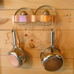 May include: Two copper pots with lids hanging on a copper rack with copper fish-shaped hooks. The rack is mounted on a wooden wall.