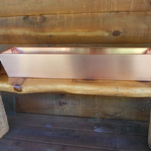 May include: A long, rectangular copper planter with slightly angled sides. The planter is sitting on a wooden bench.