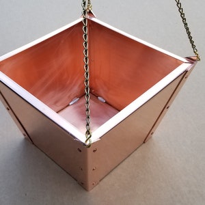 May include: A square copper hanging planter with a chain attached to each corner. The planter is empty and ready to be filled with plants.