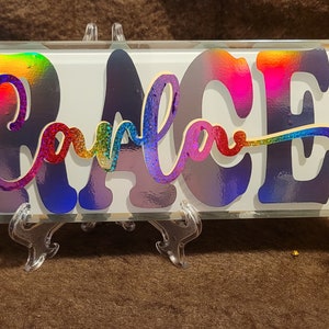May include: A glass sign with the name "GRACE" written in rainbow iridescent letters. The letters are raised and have a glittery finish.