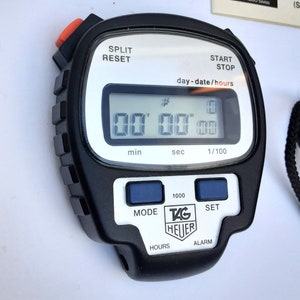 Puede incluir: Un cronómetro digital negro con una esfera plateada y una pantalla blanca. El cronómetro tiene el logotipo de TAG Heuer y las palabras "SPLIT RESET", "START STOP", "day-date/hours", "min", "sec", "1/100", "MODE", "1000", "SET", "HOURS", y "ALARM".