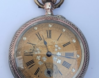 May include: Vintage pocket watch featuring a gold-coloured face with black Roman numerals and floral detailing. The watch has a gold-coloured bezel and a textured winding crown. The second hand is in a small subdial at the bottom.
