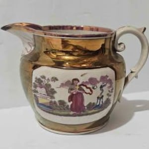 May include: A vintage ceramic pitcher with a gold metallic finish and a cream-colored central panel. The panel features a pastoral scene with figures and trees. The handle is white, and the spout is integrated into the design.