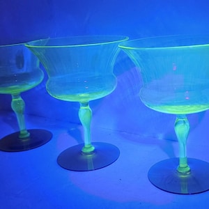 May include: Three clear glass cocktail glasses with a distinctive shape, featuring a wide bowl and a slender stem. The glasses have a neon green glow under a blue light, creating a unique visual effect. The glasses are displayed on a dark surface.