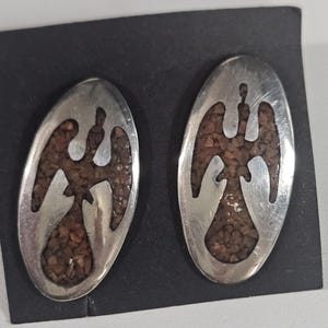 May include: A pair of oval silver earrings. Each earring has a cut-out bird design filled with small, reddish-brown stones. The earrings are presented on a black surface.