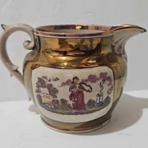 May include: Vintage ceramic pitcher with a gold-colored exterior and a cream-colored central panel. The panel features a painted scene with figures and landscape elements. The handle is a light beige color.