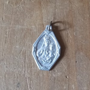 May include: Silver pendant featuring a religious image of the Virgin Mary and child. The pendant is octagonal in shape and has a loop at the top for attaching to a chain.