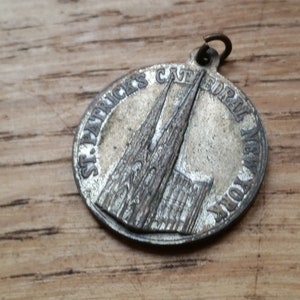 May include: A silver-toned round pendant featuring an embossed image of St. Patrick's Cathedral in New York. The text "ST. PATRICK'S CATHEDRAL NEW YORK" is inscribed around the edge. The pendant has a small loop at the top for attachment.