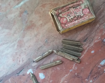 Könnte beinhalten: Eine Vintage-Schachtel mit Metallfedern für Füllfederhalter. Die Schachtel ist rot und weiß mit goldenen Lettern und den Worten "DE BAIGNOL & FARJO" auf der Vorderseite. Die Schachtel ist geöffnet und die Federn liegen auf einer Marmorfläche verstreut.