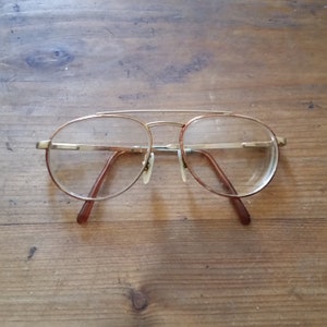 Puede incluir: Gafas redondas con montura dorada y patillas marrones. Las lentes son transparentes.