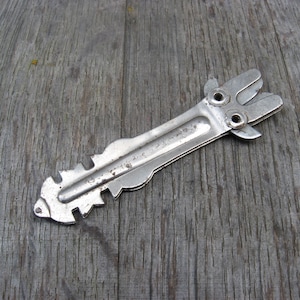 Pode incluir: Uma ferramenta de metal prateado com um design único, com várias ranhuras e uma alça com dois furos. A ferramenta está sobre uma superfície de madeira.