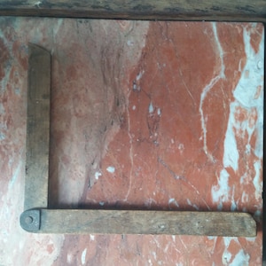 Puede incluir: Una escuadra de carpintero de madera vintage, con un diseño de ángulo recto. La herramienta está hecha de madera envejecida, mostrando signos de desgaste. La escuadra se encuentra sobre una superficie de mármol rojo y blanco.