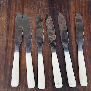 May include: Six silver butter knives with white handles. The knives have serrated edges and are arranged in a row on a wooden surface.