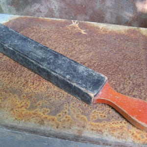 May include: A vintage straight razor strop with a black leather surface and a red wooden handle. The strop is rectangular and appears well-used, showing signs of wear. The handle is a curved shape.