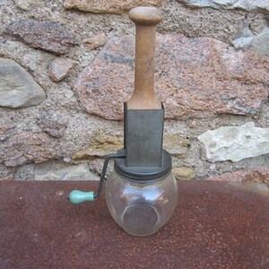May include: A vintage glass and metal food grinder with a wooden handle and a crank. The grinder has a clear glass jar with a metal lid and a metal base. The wooden handle is attached to a metal rod that goes through the lid of the jar. The crank is attached to the metal rod and is used to turn the grinder.