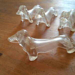 Könnte beinhalten: Fünf klare Glas-Dackel-Figuren. Die Figuren sind alle in der gleichen Pose, mit dem Kopf zur Seite geneigt und dem Schwanz eingerollt. Sie sind alle etwa 7,5 cm lang.