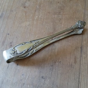 May include: Silver sugar tongs with an ornate design. The tongs are closed and have a slight curve. The tongs are on a wooden surface.