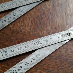 May include: Close-up of a vintage folding ruler with visible measurements in inches and centimeters. The ruler is made of metal and has black numbers printed on it. The ruler is partially open, lying on a wooden surface.