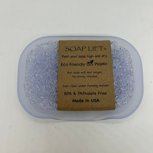May include: A clear plastic soap dish with a textured surface. The dish has a brown label that reads "SOAP LIFT", "Rest your soap high and dry", "Eco Friendly bio Plastic", "Bar soap will last longer. No messy residue.", "Just rinse under running water!", "BPA & Phthalate Free", and "Made in USA".
