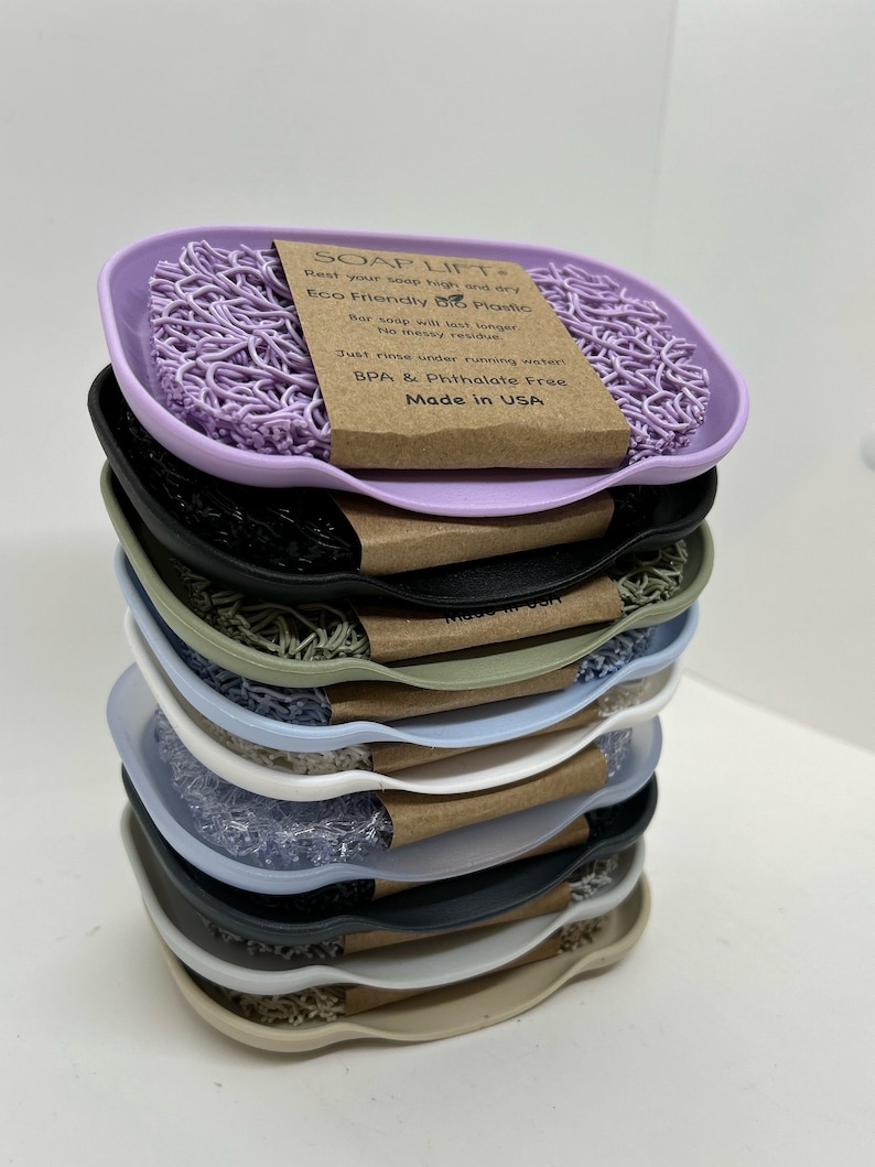 May include: A stack of eight soap dishes in various colors, including purple, black, green, white, blue, and beige. The top dish features a purple soap saver and a brown label that reads "SOAP LIFT* Rest your soap high and dry Eco Friendly Bio Plastic Bar soap will last longer. No messy residue Just rinse under running water BPA & Phthalate Free Made in USA"