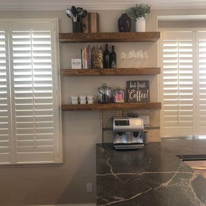 May include: Three horizontal wooden shelves mounted on a wall, holding kitchen items including a coffee machine, mugs, wine glasses, bottles, a jar of corks, and a sign that reads but first. Coffee! and another sign that reads BLESS.