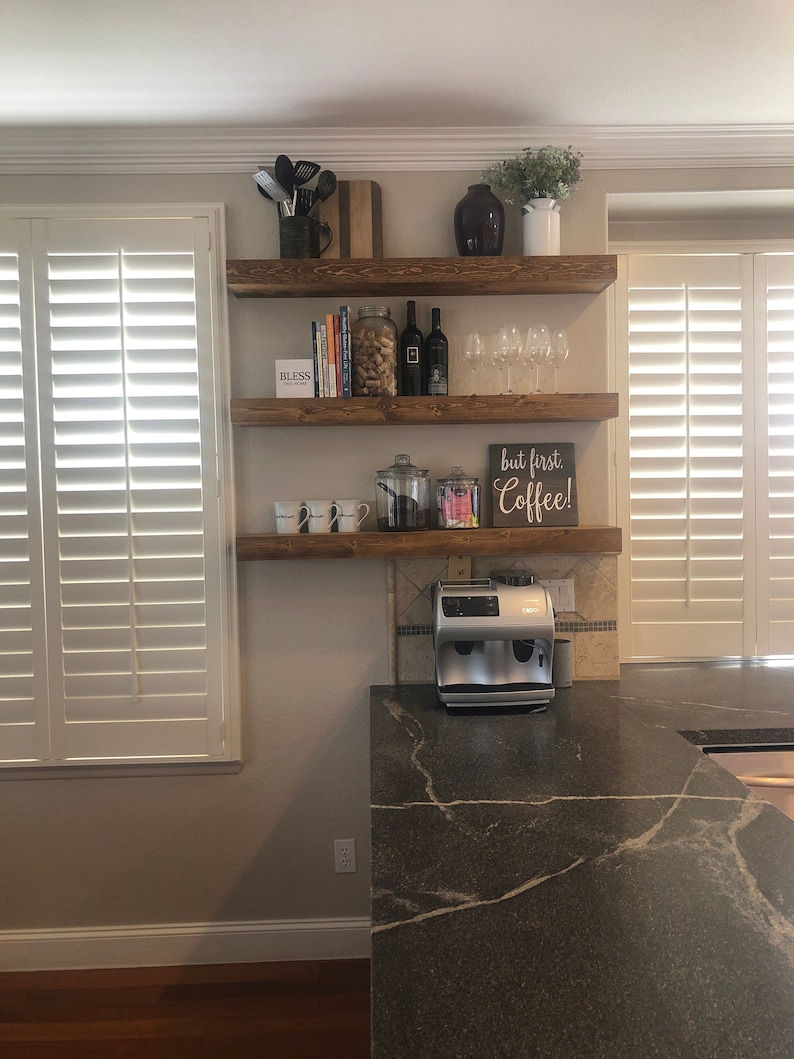 May include: Three horizontal wooden shelves mounted on a wall, holding kitchen items including a coffee machine, mugs, wine glasses, bottles, a jar of corks, and a sign that reads but first. Coffee! and another sign that reads BLESS.