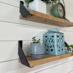 May include: Two wooden shelves with black metal brackets. The top shelf has a white planter with a green plant and a teal clock. The bottom shelf has a gray planter with a green plant, a teal lantern, and a small white planter with a green plant.