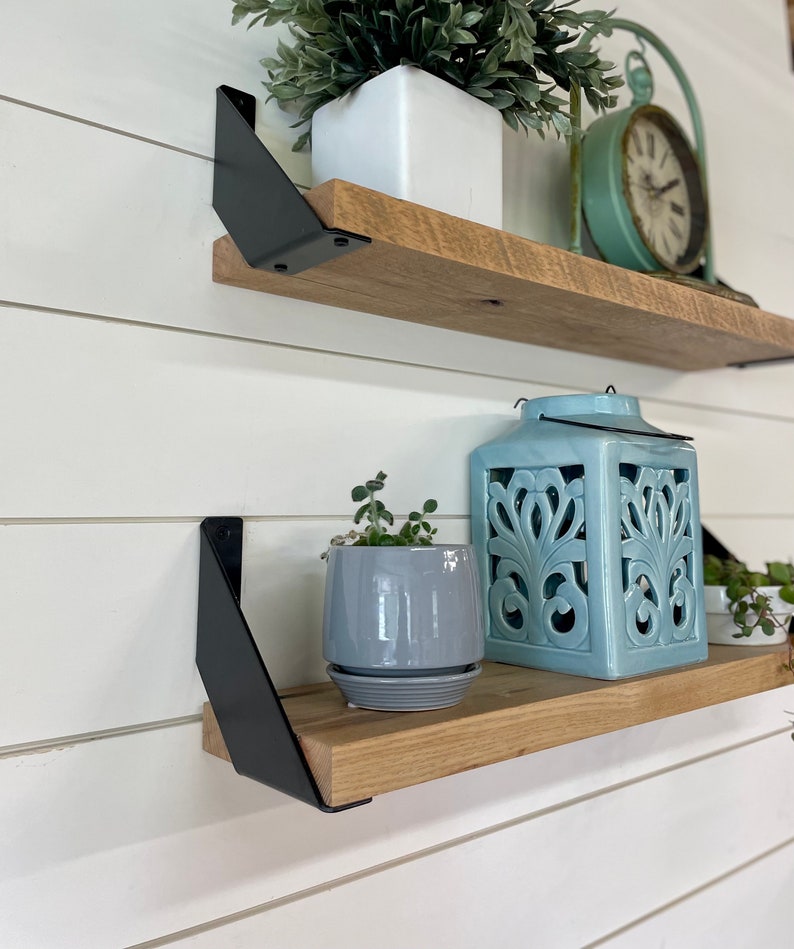 May include: Two wooden shelves with black metal brackets. The top shelf has a white planter with a green plant and a teal clock. The bottom shelf has a gray planter with a green plant, a teal lantern, and a small white planter with a green plant.