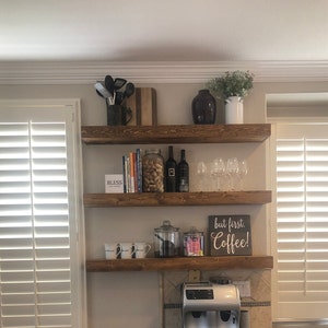 May include: Three wooden shelves mounted on a wall, with various items on them, including books, wine glasses, a coffee maker, and a sign that says "but first, coffee!"