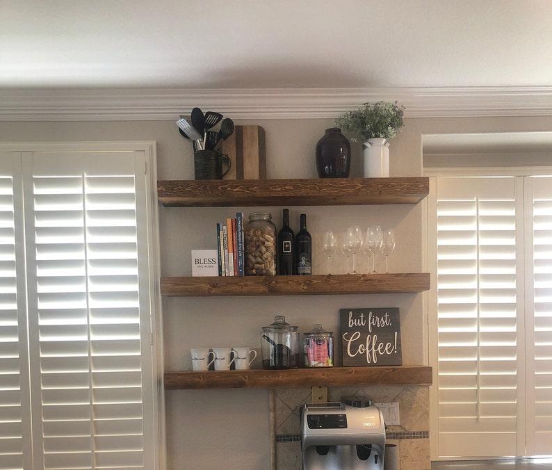 May include: Three wooden shelves mounted on a wall, with various items on them, including books, wine glasses, a coffee maker, and a sign that says "but first, coffee!"