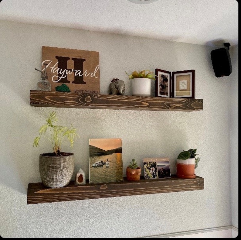 May include: Two wooden shelves with a brown stain, mounted on a white wall. The top shelf has a wooden sign with the text "Hayward EST 2020" and a small potted plant. The bottom shelf has a framed photo, a small potted plant, and a book.