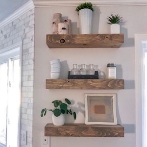 May include: Three wooden shelves mounted on a white wall, with various decorative items on each shelf, including plants, vases, and candles.