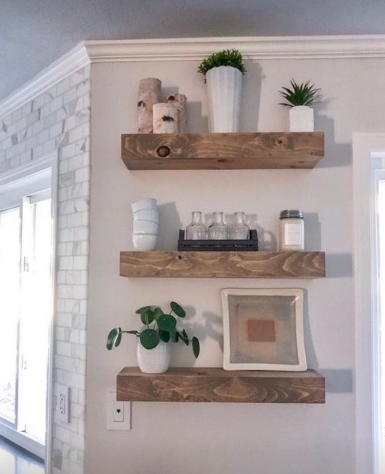 May include: Three wooden shelves mounted on a white wall, with various decorative items on each shelf, including plants, vases, and candles.