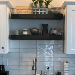 May include: Two dark brown wooden floating shelves mounted between white cabinets. The top shelf holds a wooden sign reading What Happens At Gigi's House Stays at Gigi's House, a metal crate with flowers, and a bowl. The bottom shelf holds mugs, a teapot, a pitcher, and a small framed picture.
