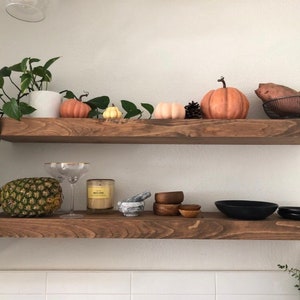 May include: Two horizontal wooden shelves mounted on a wall. The top shelf holds a potted plant, three decorative pumpkins, a pinecone, and a sweet potato in a wire basket. The bottom shelf holds a pineapple, two glass coupes, a candle, a marble mortar and pestle, three small wooden bowls, and two black bowls.