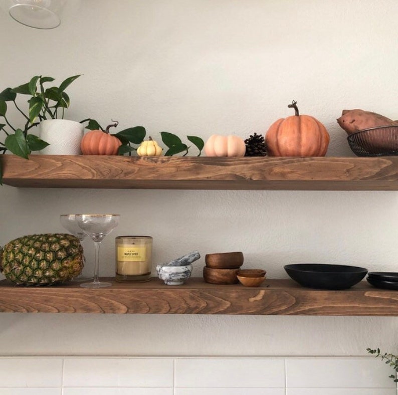 May include: Two horizontal wooden shelves mounted on a wall. The top shelf holds a potted plant, three decorative pumpkins, a pinecone, and a sweet potato in a wire basket. The bottom shelf holds a pineapple, two glass coupes, a candle, a marble mortar and pestle, three small wooden bowls, and two black bowls.