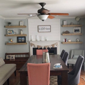 May include: A dining room with a wooden table and chairs. The walls are painted a light gray and there are three wooden shelves on each wall. The shelves are decorated with various items, including books, plants, and pictures. The text "be still & know" is written on the wall above the fireplace.