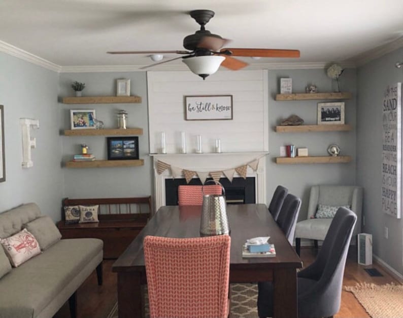 May include: A dining room with a wooden table and chairs. The walls are painted a light gray and there are three wooden shelves on each wall. The shelves are decorated with various items, including books, plants, and pictures. The text "be still & know" is written on the wall above the fireplace.