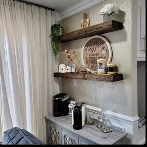 May include: Two wooden shelves with a coffee themed sign that reads "Coffee is always a good idea". The shelves are mounted on a wall with a light brown textured wallpaper. There are various coffee related items on the shelves, including jars, a coffee maker, and a coffee pod machine.