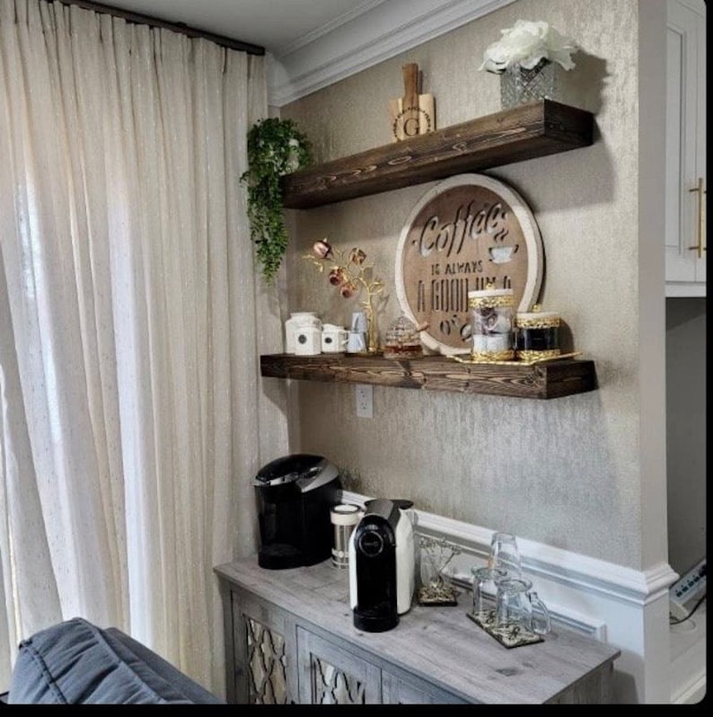 May include: Two wooden shelves with a coffee themed sign that reads "Coffee is always a good idea". The shelves are mounted on a wall with a light brown textured wallpaper. There are various coffee related items on the shelves, including jars, a coffee maker, and a coffee pod machine.