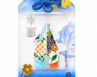 May include: A rectangular blue and clear plastic omamori charm featuring a central origami crane with colorful floral patterns. The charm includes a blue cord, gold-colored leaf accents, and Japanese text characters for good luck printed on the front.