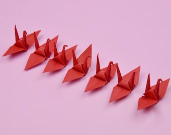 May include: A row of eight red origami cranes arranged in a line on a pink background.