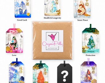 May include: A collection of eight rectangular resin omamori charms featuring colorful origami paper inserts and Japanese kanji, arranged around a central brown envelope with an Origami Polly logo featuring a pink paper crane.