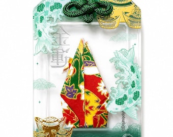 May include: A rectangular green omamori charm featuring a clear center window displaying a folded paper crane with red, green, and yellow floral patterns. The charm includes gold-colored ingot accents, koi fish illustrations, and the text WEALTH.