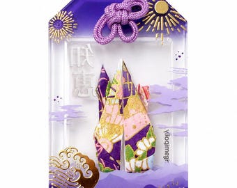 May include: A rectangular purple omamori charm featuring a clear center window displaying a folded paper crane with floral patterns. The charm is decorated with gold foil fireworks, waves, and the word WISDOM printed at the bottom.