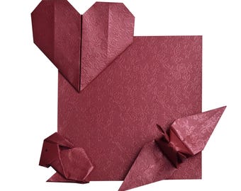 May include: A burgundy origami heart, sheep, and crane are arranged on a textured square. The paper has a subtle floral pattern. The heart is at the top, the sheep at the bottom left, and the crane at the bottom right.